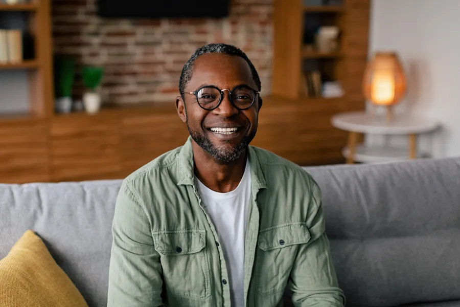 Andropause-Doctor A middle aged black man in a green button up shirt sits on his couch smiling, happy with his treatment for andropause and low testosterone from Brandi Terra, NP at Oak & Stone Wellness in East San Diego.