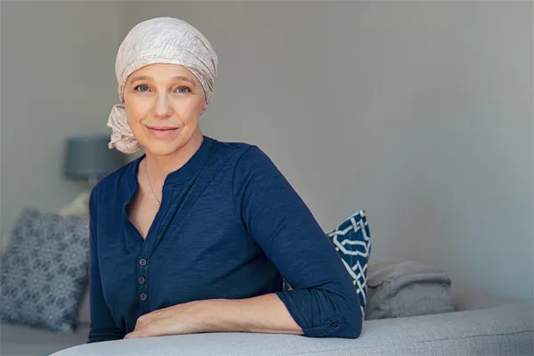 Functional-Oncology-Doctor A woman wearing a blue shirt with a white scarf tied around her head sitting on a couch and smiling at the camera, representing sensitive functional oncology treatment from Brandi Terra, NP at Oak & Stone Wellness in East San Diego.