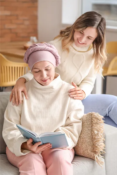 Functional-Oncology-Treatment A woman with a scarf tied around her head sits on a couch reading while another woman sits behind her with her hands on her shoulders warmly, representing sensitive functional oncology treatment from Brandi Terra, NP at Oak & Stone Wellness in East San Diego.