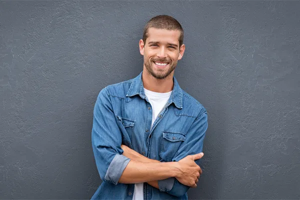 Hormones-for-Men-Doctor A man in a denim shirt stands smiling against a gray-blue wall, pleased with his testosterone hormone therapy from Brandi Terra, NP at Oak & Stone Wellness in East San Diego.