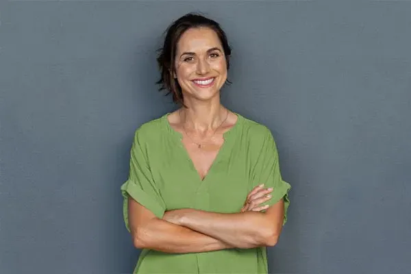Hormones-for-Women-Doctor A woman in a green shirt stands smiling against a gray-blue wall, pleased with her hormone therapy from Brandi Terra, NP at Oak & Stone Wellness in East San Diego.