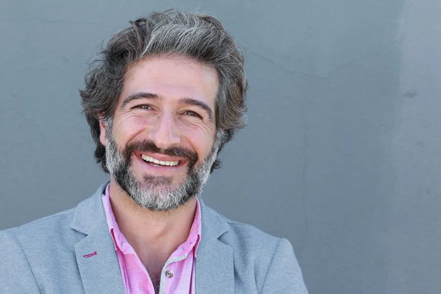 Hypogonadism-Clinic A smiling man with salt and pepper hair and beard wearing a pink shirt and gray blazer stands agains a gray wall. Get hypogonadism treatment from Brandi Terra, NP at Oak & Stone Wellness in East San Diego.