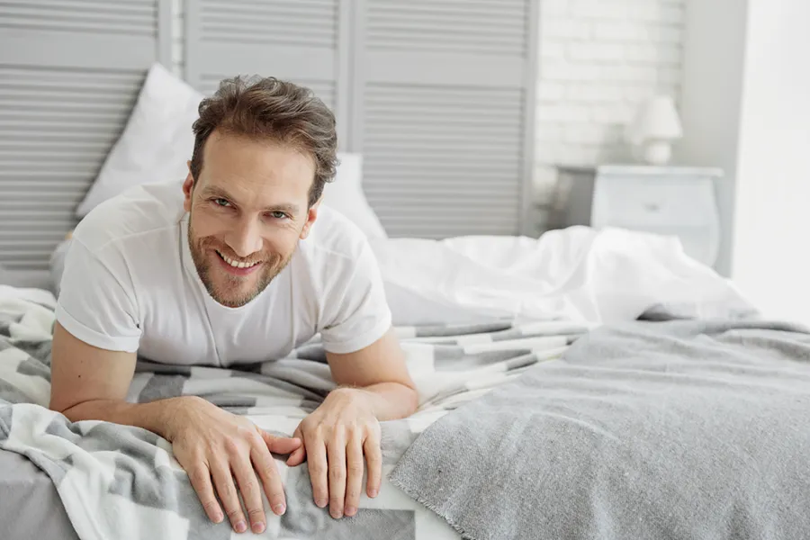 Low-Libido-in-Men-Doctor A dirty-blonde haired man lays on his stomach in bed smiling. Schedule treatment for low libido in men from Brandi Terra, NP at Oak & Stone Wellness in East San Diego.