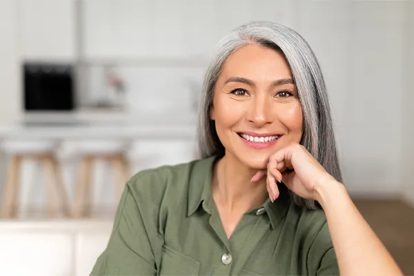 Menopause-Clinic An asian woman with long gray hair and a green shirt sitting and smiling on her couch, happy with her menopause care from [NAME} in East San Diego.