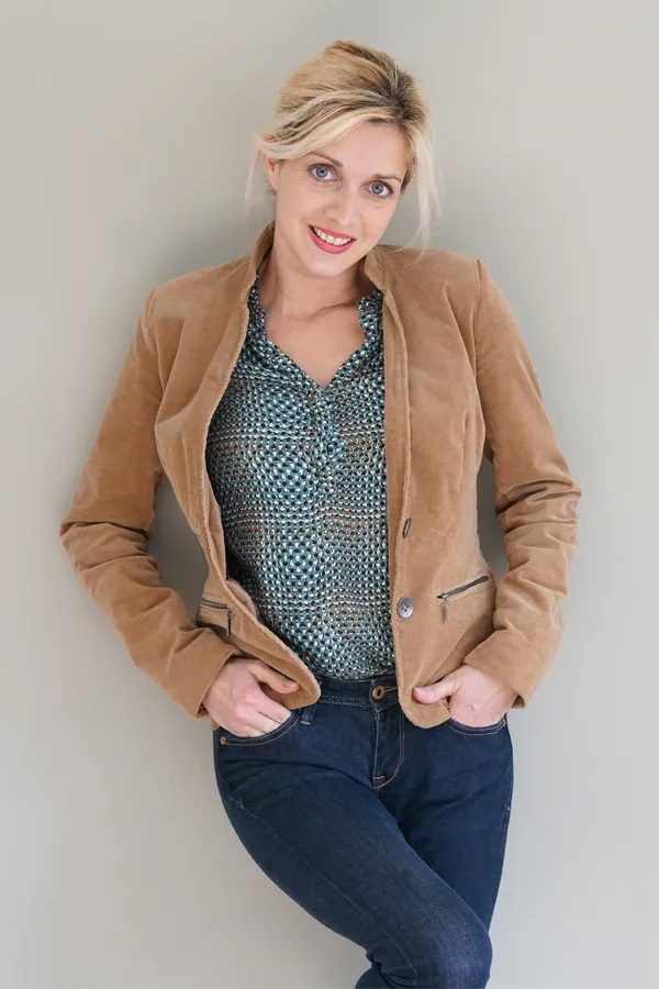 Metabolic-Syndrome-Treatment A smiling blonde woman in a tan leather jacket leans against a gray wall. Schedule metabolic syndrome from Brandi Terra, NP at Oak & Stone Wellness in East San Diego.