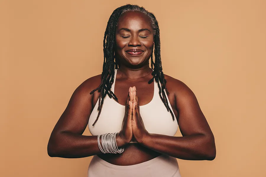 Obesity-Clinic An older African American woman confidently posing against a warm background smiling with hands in a prayer pose. Brandi Terra, NP at Oak & Stone Wellness treats obesity using functional medicine in East San Diego.