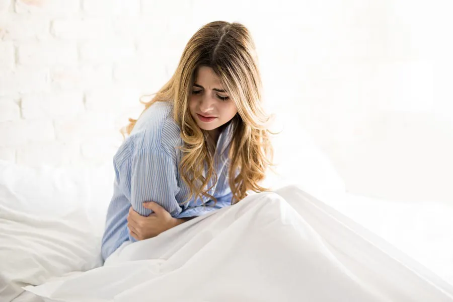 PMS-Therapy A woman with long blonde hair sits in her bed holding her stomach in pain, considering getting care for PMS from Brandi Terra, NP at Oak & Stone Wellness in East San Diego.