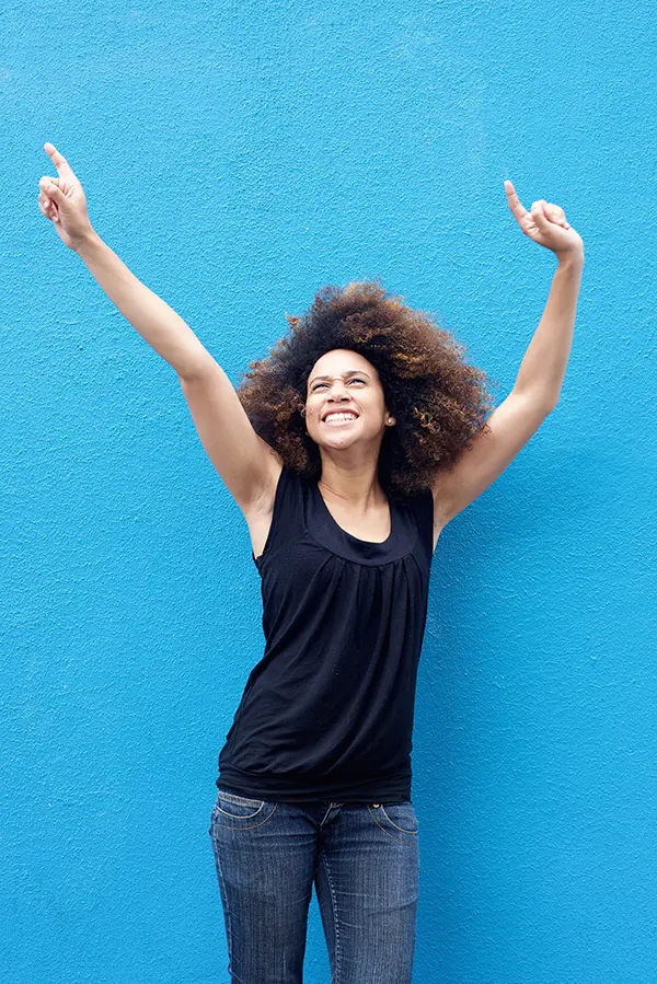 PMS-Treatment A woman in a dark blue tank top standing in front of a bright blue wall, raising her arms in celebration of relief from PMS from Brandi Terra, NP at Oak & Stone Wellness in East San Diego.