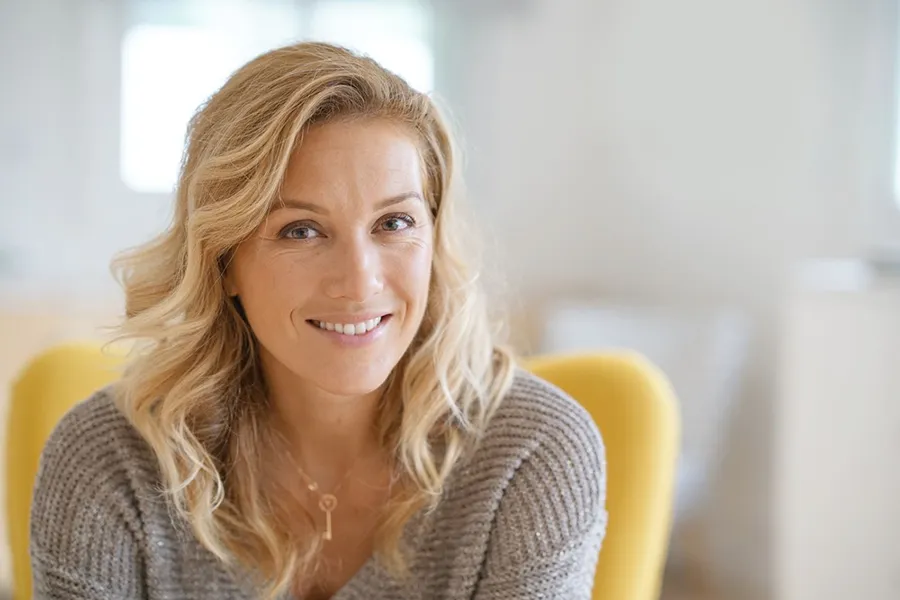 Perimenopause-Doctor A blonde woman wearing a a gray sweater sits inside in a yellow chair, smiling and happy with her perimenopause treatment from Brandi Terra, NP at Oak & Stone Wellness in East San Diego.