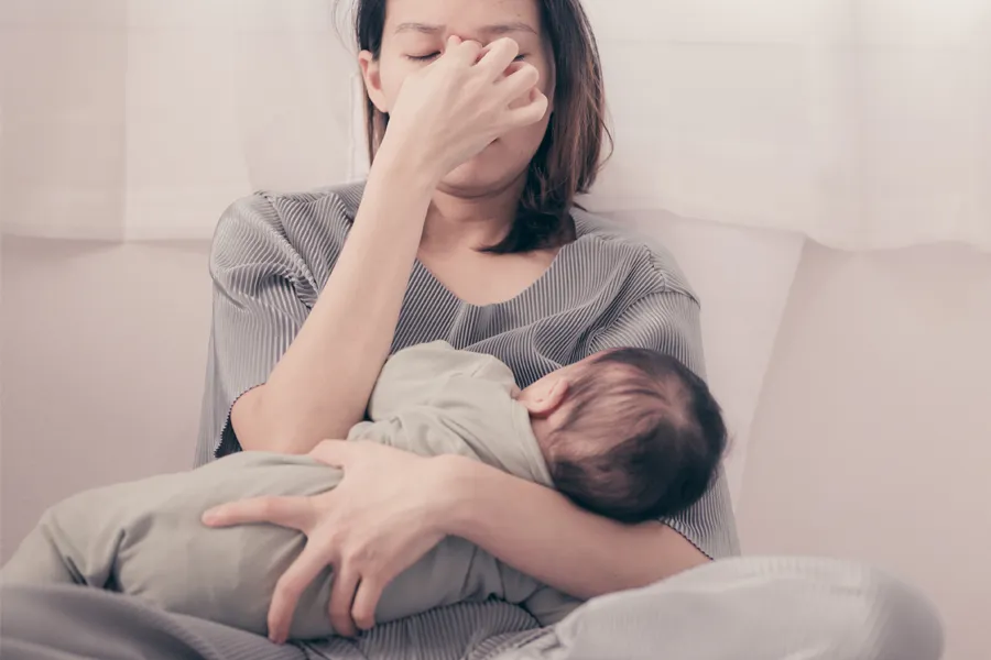 Postpartum-Hormone-Imbalance-Clinic A woman nursing an infant on couch rubs her eyes with exhaustion. Get treatment for postpartum hormone imbalance from Brandi Terra, NP at Oak & Stone Wellness in East San Diego.