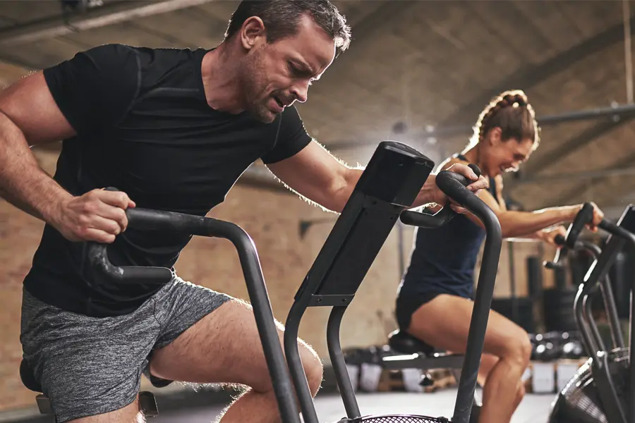 Tirzepatide-Doctor A couple in a brick gym work as hard as they can on exercise bikes, inspired by successful Tirzepatide treatment from Brandi Terra, NP at Oak & Stone Wellness in East San Diego.