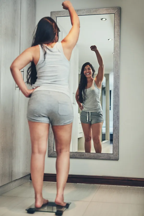 Tirzepatide-Treatment A woman in workout clothes stands on a scale looking in the mirror and pumping her hands in celebration of her successful Tirzepatide treatment from Brandi Terra, NP at Oak & Stone Wellness in East San Diego.
